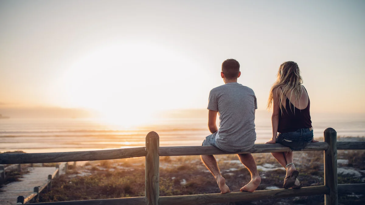 Two people sitting on a railing on beach looking out into the water.